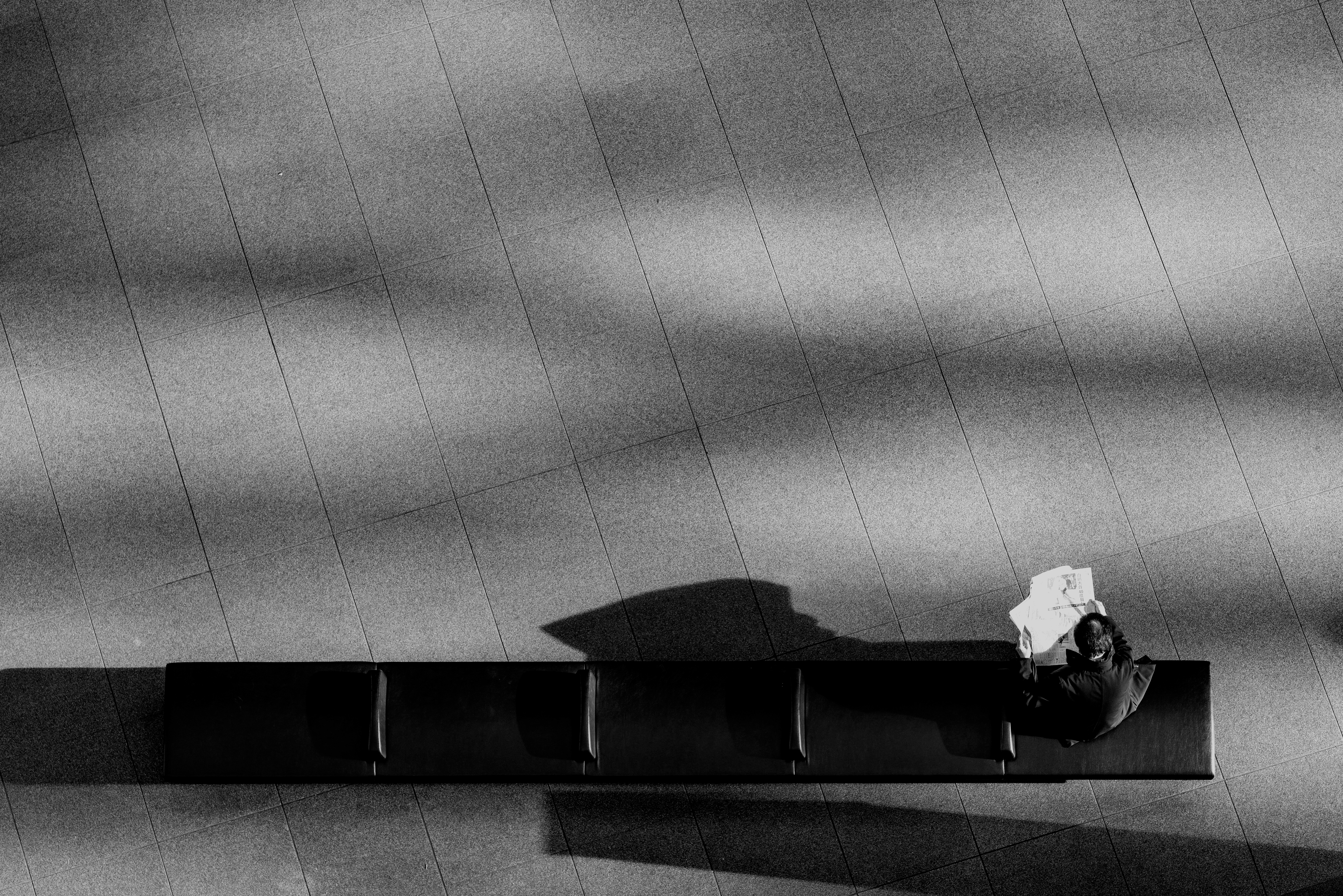 Overhead view of a man reading a newspaper alone on a long bench in a sunlit plaza, black and white